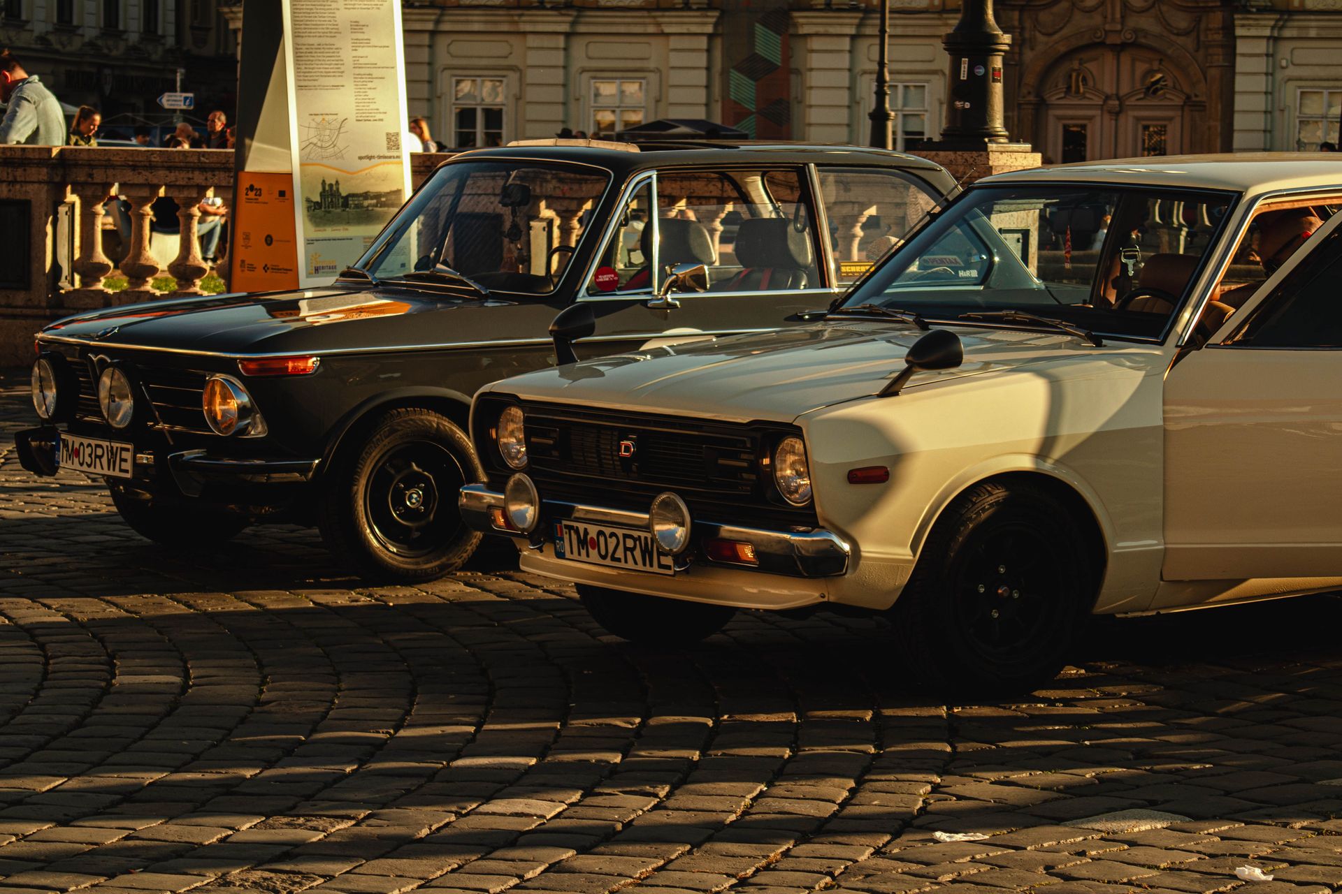Two vintage cars parked on a cobblestone street in a historic European town during the day.